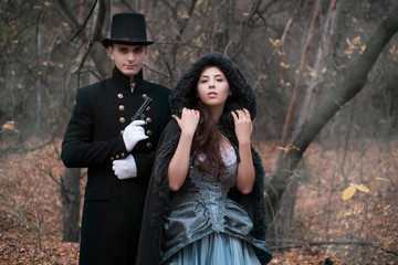 A man in a nineteenth century suit holds a gun, protects a girl. Young man and woman in historical costumes in a gloomy forest