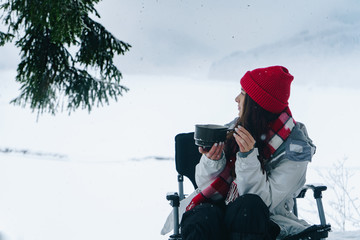 Beautiful woman sitting near fire with a cup of tea in the winter forest with scenic mountain view. Tourist camp with campfire. Camping and hiking, winter adventure, nature landscape. Side view