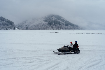 Side view of man and woman riding fast on a snowmobile on the frozen lake in the mountains with the scenic view.