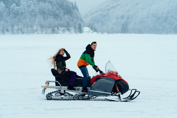Fototapeta na wymiar Man and woman riding on a snowmobile on the frozen lake in the mountains with the scenic view. Pine trees covered with snow. Side view