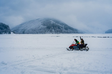 Fototapeta na wymiar Man and woman riding on a snowmobile on the frozen lake in the mountains with the scenic view. Pine trees covered with snow. Side view