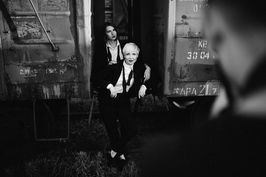 Couple of woman are posing in black classic suit at the rust background at railway station at the sunset