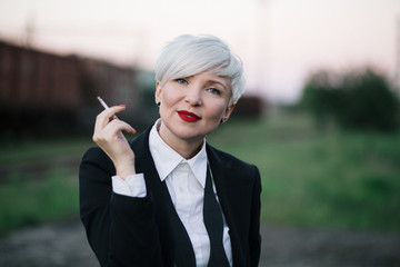 Stylish blonde haired woman in black tie suit smoking at the railway station