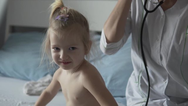 A doctor in the hospital is listening to a little girl with a stetus. The baby is crumpled, the woman in the white coat smoothes the baby on the back. Pediatrician examination at home