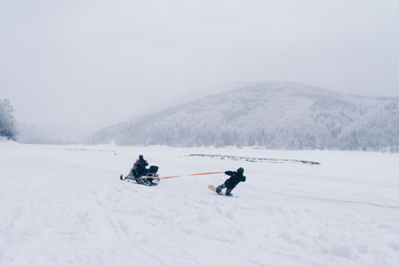 Snowboarder man riding on a snowmobile holding a rope. On the frozen lake between beautiful snowy forest. Extreme sport