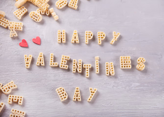 Text Valentines day written with macaroni in the form of letters and two red hearts cookies on grey wooden table background