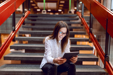 Smiling female student with eyeglasses and brown hair using tablet while sitting on the stairs.