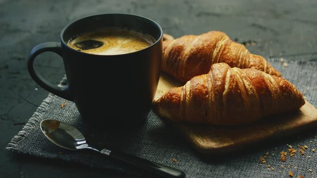 From above view of two fresh croissants and black mug with coffee placed on napkin on gray background of table