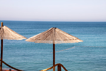 Lonely wicker sun umbrella at mediterranean beach by sea. Natural bamboo sunshades and summer umbrella parasol on ocean beach. Large straw beach umbrellas against blue water and sky. Vacation, travel.