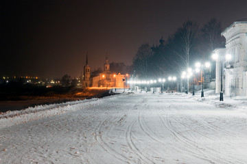 The embankment of Veliky Ustyug on a winter night: a snowy road lit by lanterns and a church against a dark sky