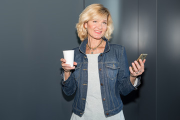 Mature woman with a smartphone in her hands and a glass of hot coffee on a gray background.