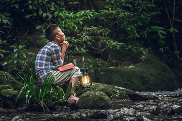 Boy reading book or bible and praying with oil lamp at river. christian religion concept.