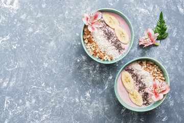 Healthy breakfast of yogurt with banana, chia seeds, coconut flakes and almonds on a gray background, decorated with pink alstroemeria flowers. Top view, copy space.