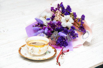 A bouquet of dried flowers in blue tones, a cup of coffee on a saucer, red glasses on a light wooden background.