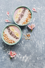 Fresh yogurt with banana, chia seeds, coconut flakes and almonds on a gray background, decorated with pink alstroemeria flowers. Top view, copy space. The concept of healthy eating.
