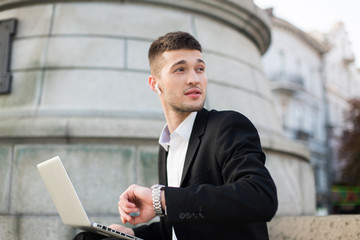 Young pensive businessman in black suit with wireless earphones dreamily looking aside with hand watch and laptop on knees outdoor