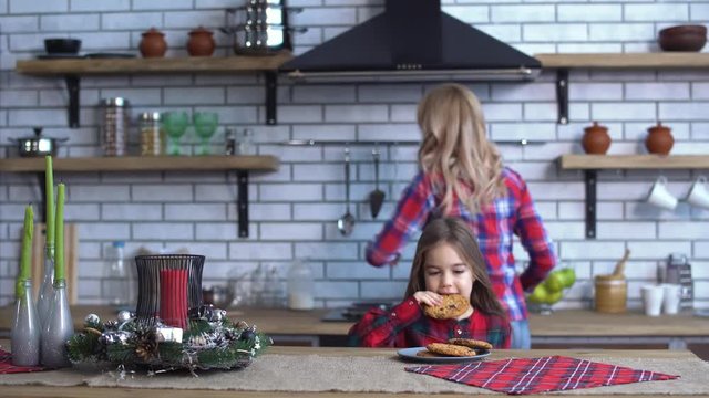 Young mother in plaid shirt with long blond hair cooking breakfast and small naughty girl bite one cookie
