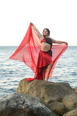 A brunette woman in a red dress with a shawl on a rock on the seashore is dancing oriental.