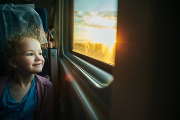 Beautiful little girl looking out train window outside, while it moving. Going on vacations and traveling by railway in summer. Sunset time.
