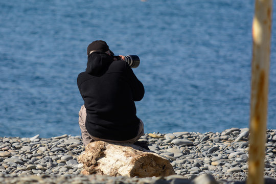 Photographer in dark clothes with camera with big lens is sitting on the beach on the rocks and taking pictures
