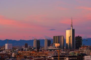 Fototapeta premium Panoramę Mediolanu o zachodzie słońca z nowoczesnymi drapaczami chmur w dzielnicy biznesowej Porta Nuova we Włoszech. Panoramiczny widok na miasto Mediolan. W tle pasmo górskie Alp Lombardii.
