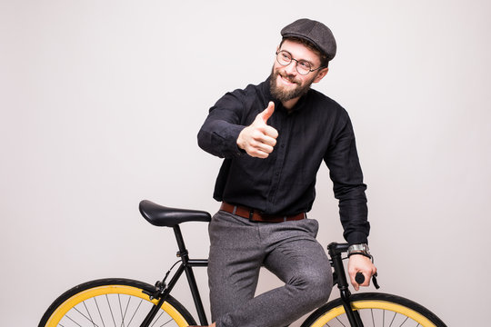 Portrait of a smiling man wearing in eyeglasses and hat leaning on bicycle with thumbs up gesture over white background