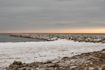 Sunset in cold winter day at the snowy seashore and rocky mole.