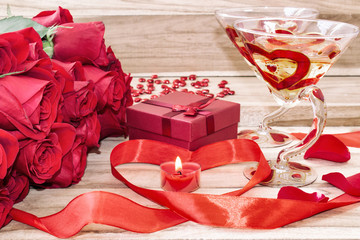 Festive background to the Valentine's day. A bouquet of red roses, a gift box, a heart-shaped candle and a red ribbon with a heart and two glasses painted for champagne. On a wooden background.