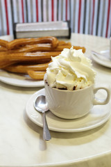 A typical spanish merienda, light meal: a cup of hot chocolate with milk cream on top of it, in front of typical Spanish fried churros