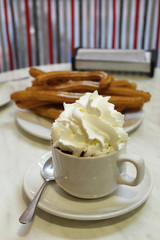 A typical spanish merienda, light meal: a cup of hot chocolate with milk cream on top of it, in front of typical Spanish fried churros