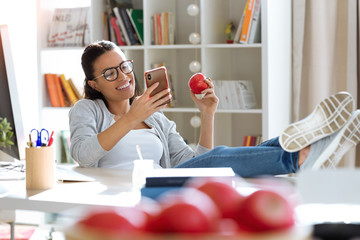 Pretty young business woman using her mobile phone while eating red apple in the office.