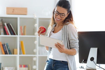 Pretty young business woman using her digital tablet while eating red apple in the office.