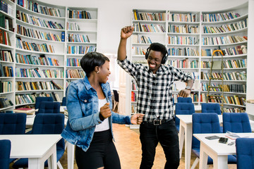 Beautiful African-American cheerful couple of students have a fun and dancing in the library. The girl smiles and the boy dances while listening to music. Happy couple smiling and dancing in library
