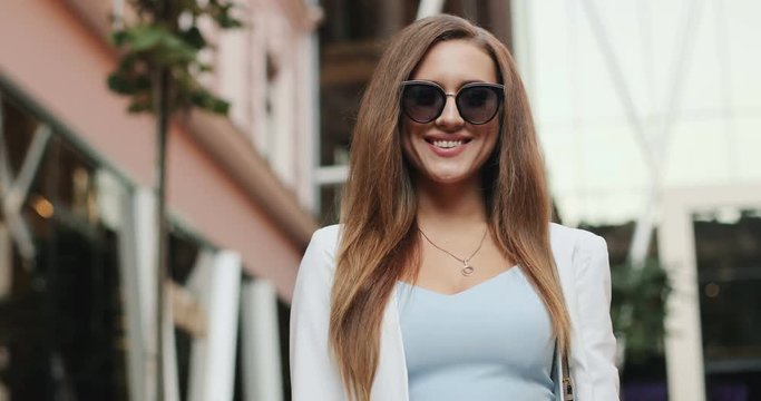 Portrait of teh pretty young Caucasian woman in sunglasses and stylish outfit smiling to the camera outdoors in the nice city.