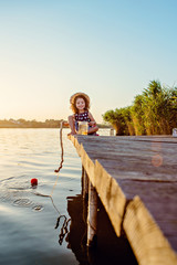 Beautiful girl in a hat at sunset catches fish on the pier at the lake.