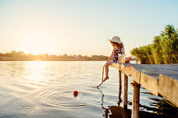 Beautiful girl in a hat at sunset catches fish on the pier at the lake.