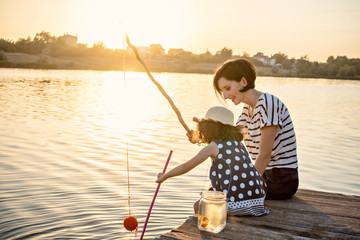 Mom and her little daughter are fishing with a fishing rod on a pier at sunset by the lake.