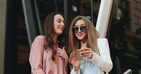 Portrait shot of the two stylish beautiful young female best friends reading something on the smartphone and they laughing. Outdoor.