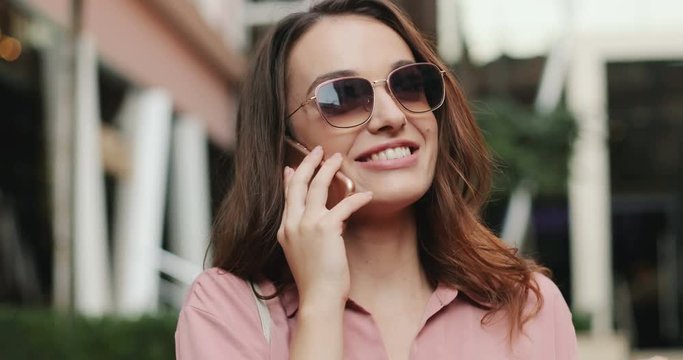 Close up of the young pretty Caucasian woman with dark hair and in sunglasses speaking on the mobile phone and smiling outside.