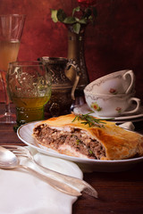 Multi-layer pie on an oval plate. A glass, cups, a jug, a glass, a vase, a nickel silver spoon, drapery. Viennese pie