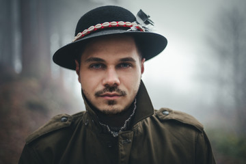 Portrait of male model with hat and beautiful eyes in deep forest at twilight. Woodsman in misty and foggy forest looking to the camera. Close up portrait of fashionable man.