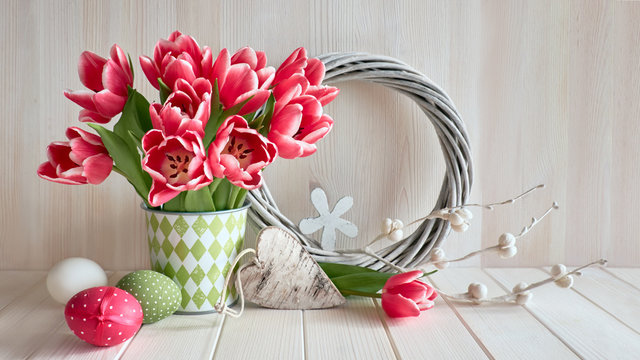 Springtime background with pink stripy tulups, Easter eggs, wooden heart and wattled wreath on light wood