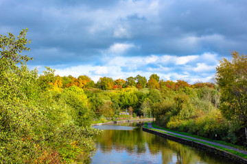 Fototapeta premium Trent and Mersey canal in autumn.Moody sky above woodland and trees with leaves changing colour growing on banks of canal.Idyllic scenery of rural Britain.
