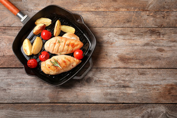 Grill pan with fried chicken breast and garnish on wooden background, top view. Space for text