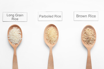 Spoons with different types of uncooked rice and cards on white background, top view
