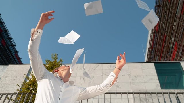 SLOW MOTION BOTTOM UP CLOSE UP: Young businessman throws paperwork in the sky after quitting his job. Work documents falling from the blue sky as cheerful man celebrates finishing a difficult project.