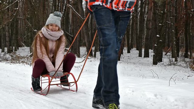 Teenager girl enjoying sleigh ride. Young people sledge in snowy forest in winter. Boy pulling sled - slow motion