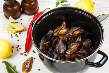 Spicy mussels in black saucepan. White wooden background decorated with lemon, hot peppers and greens