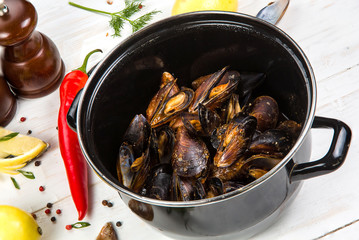 Spicy mussels in black saucepan. White wooden background decorated with lemon, hot peppers and greens