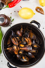 Spicy mussels in black saucepan. White wooden background decorated with lemon, hot peppers and greens